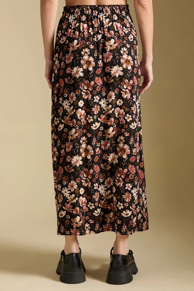 A model is shown from the back wearing a black midi skirt with a dense floral pattern in pink, beige, and white, paired with black platform shoes against a solid beige background.
