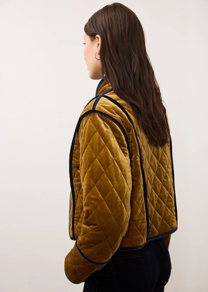 A woman shown from the side and back wearing a mustard yellow quilted velvet jacket with dark navy piping, set against a plain off-white background.