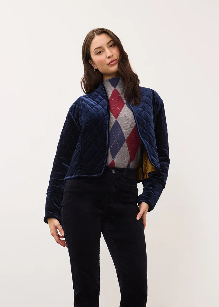 A woman models a navy blue quilted zip-up jacket over a multicolor argyle sweater, paired with dark trousers and brown ankle boots, standing against a plain off-white wall.