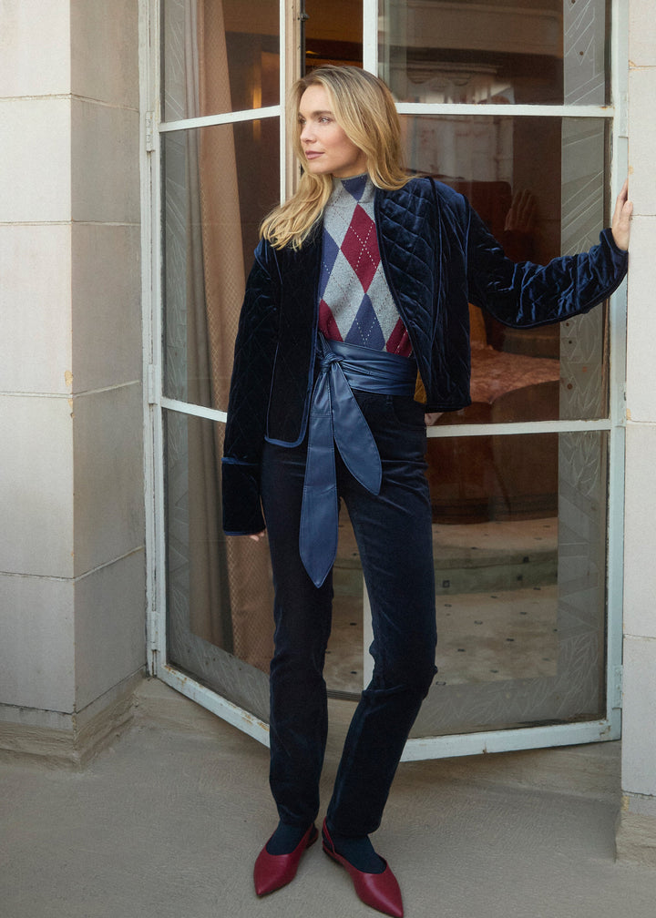 A woman stands in front of a glass-paneled doorway wearing a dark navy velvet jacket and matching trousers, a blue waist tie, a red and gray argyle sweater vest, and dark red pointed shoes.