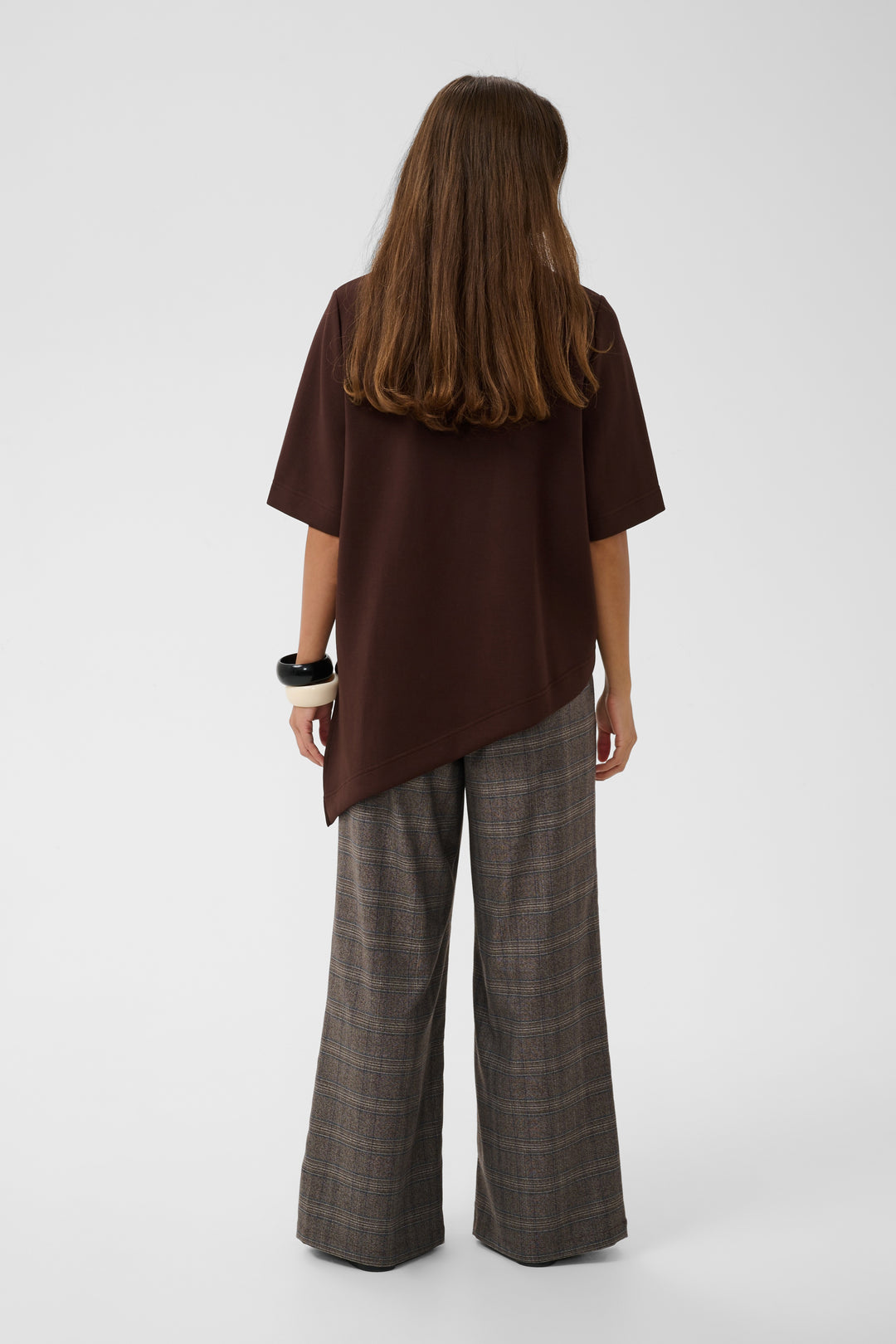 A woman shown from the back wearing a dark brown short-sleeve top with an asymmetrical hem and wide-leg plaid trousers in brown and gray tones, photographed against a light gray studio background.