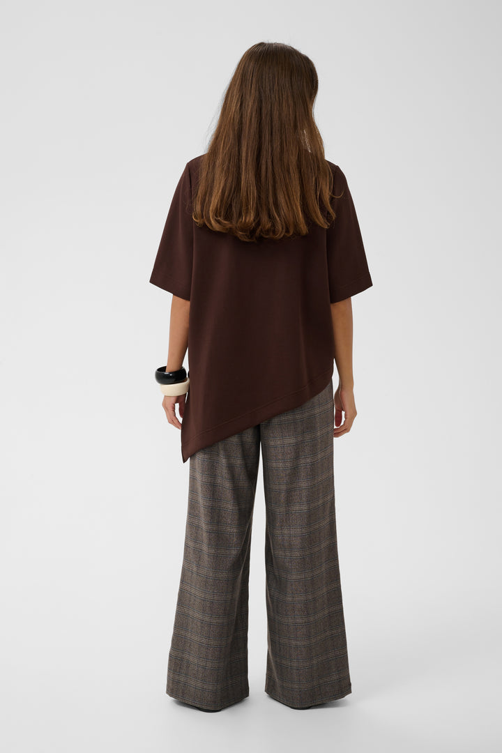 A woman shown from the back wearing a dark brown short-sleeve top with an asymmetrical hem and wide-leg plaid trousers in brown and gray tones, photographed against a light gray studio background.