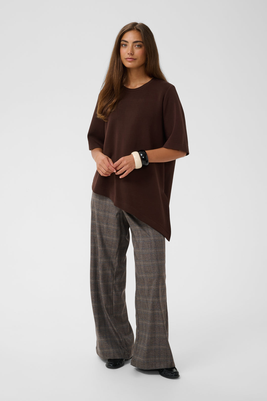 A woman modeling an oversized dark brown short-sleeve top with an asymmetrical hem, paired with brown and gray plaid wide-leg trousers, against a light gray studio background.