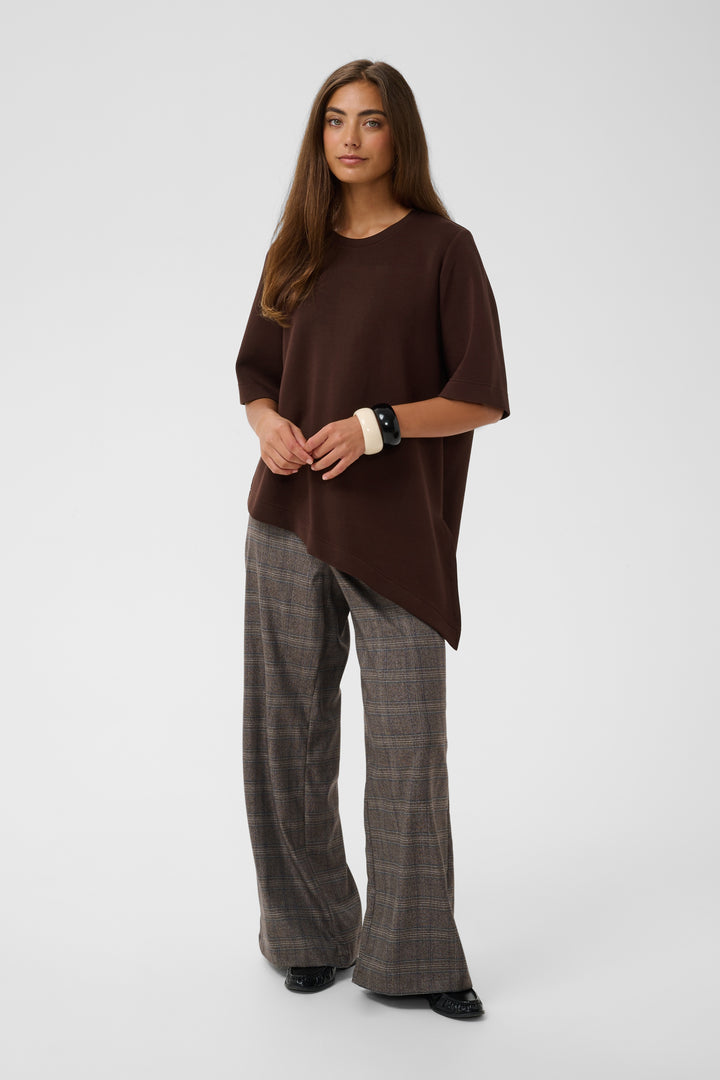 A woman modeling an oversized dark brown short-sleeve top with an asymmetrical hem, paired with brown and gray plaid wide-leg trousers, against a light gray studio background.