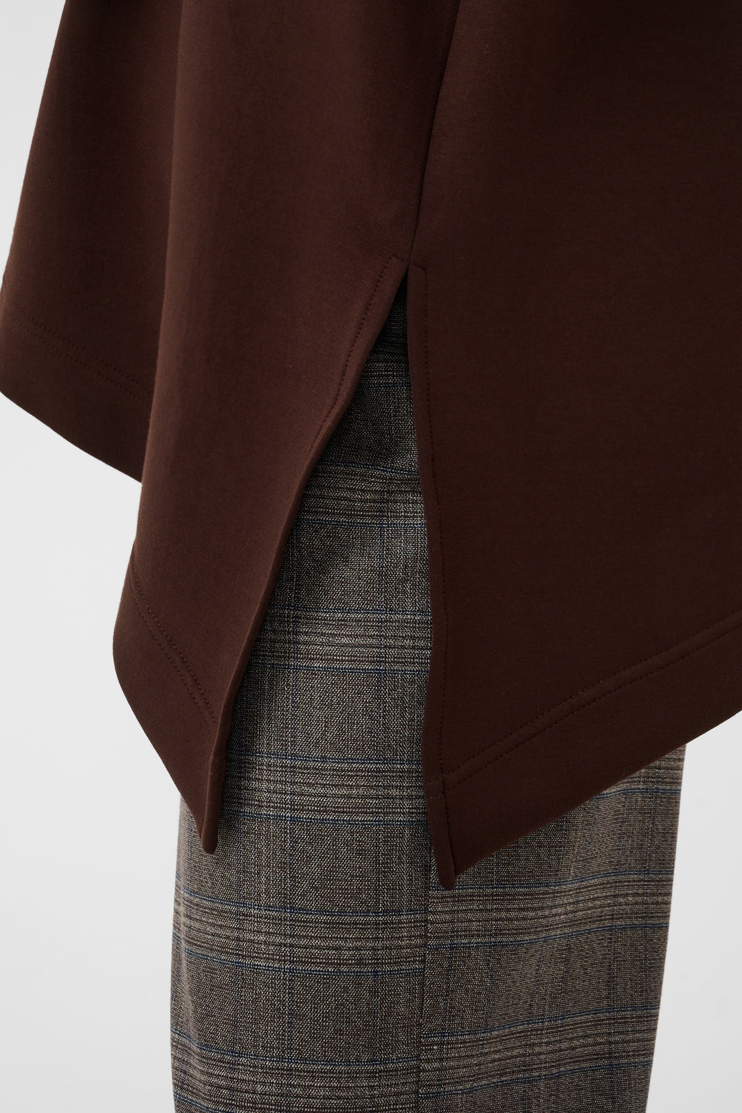Close-up of a dark brown draped garment with a side slit, layered over gray and brown plaid trousers, against a plain light background.