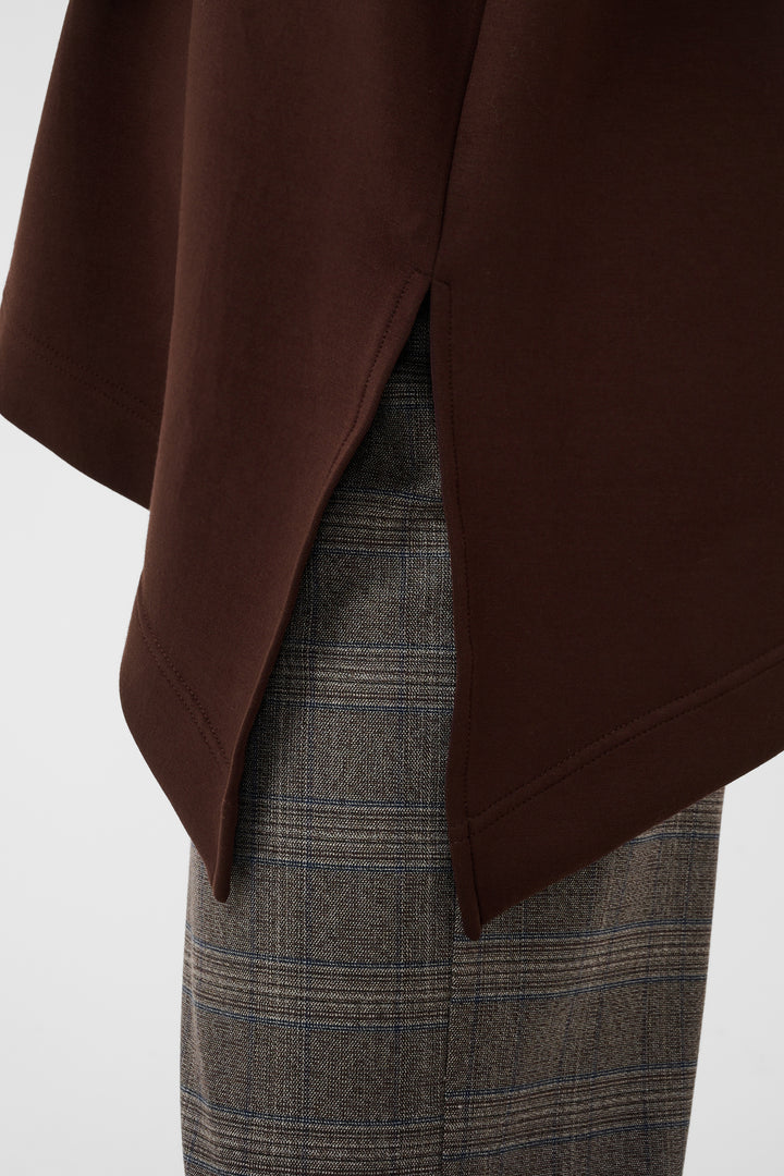 Close-up of a dark brown draped garment with a side slit, layered over gray and brown plaid trousers, against a plain light background.