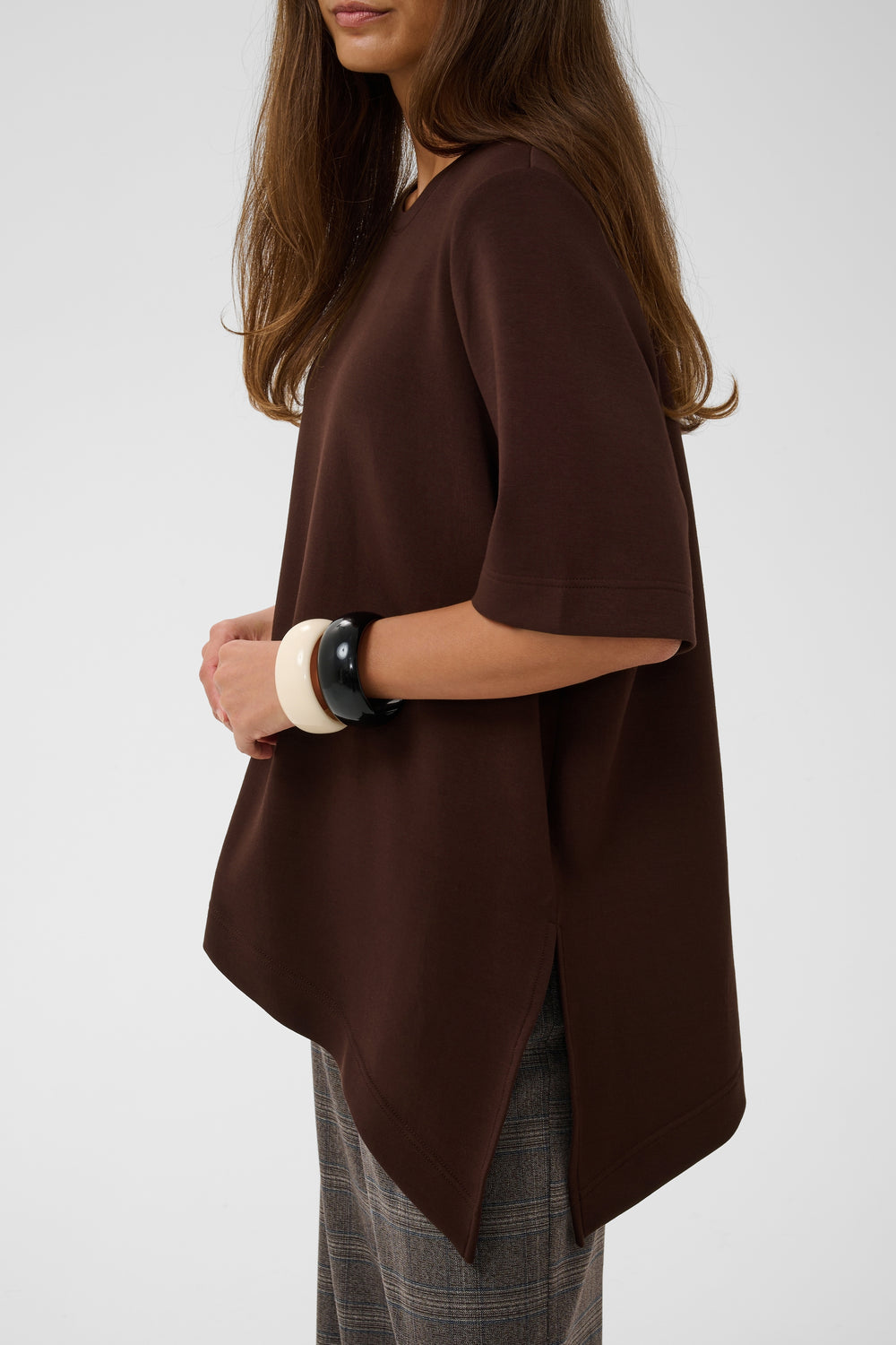A woman wearing a dark brown short-sleeve top with an asymmetrical hem, paired with gray plaid trousers, shown from a side profile against a light gray background.