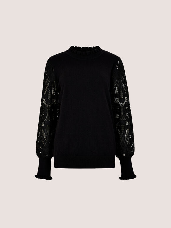 A black knit sweater with a ruffled mock neck and sheer, patterned lace sleeves, displayed against a solid light beige background.