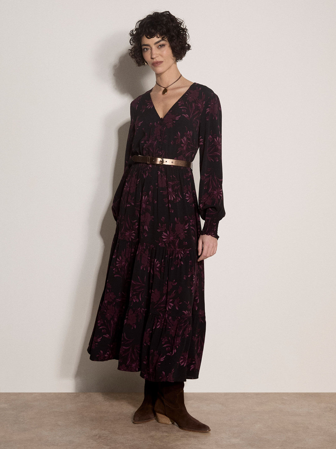 A woman models a long-sleeved midi dress with a dark floral pattern, a V-neckline, and a thin metallic gold belt at the waist. She is standing against a plain off-white wall.