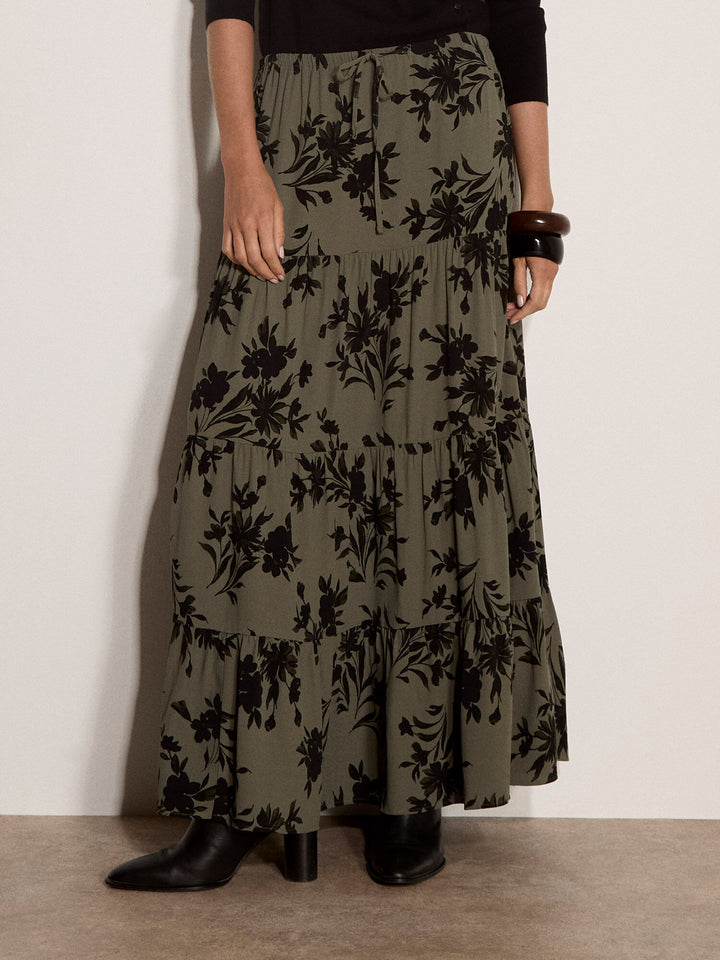 A model wearing a long, tiered maxi skirt in olive green with a black floral print, paired with black ankle boots and a dark top, standing against a plain off-white wall.