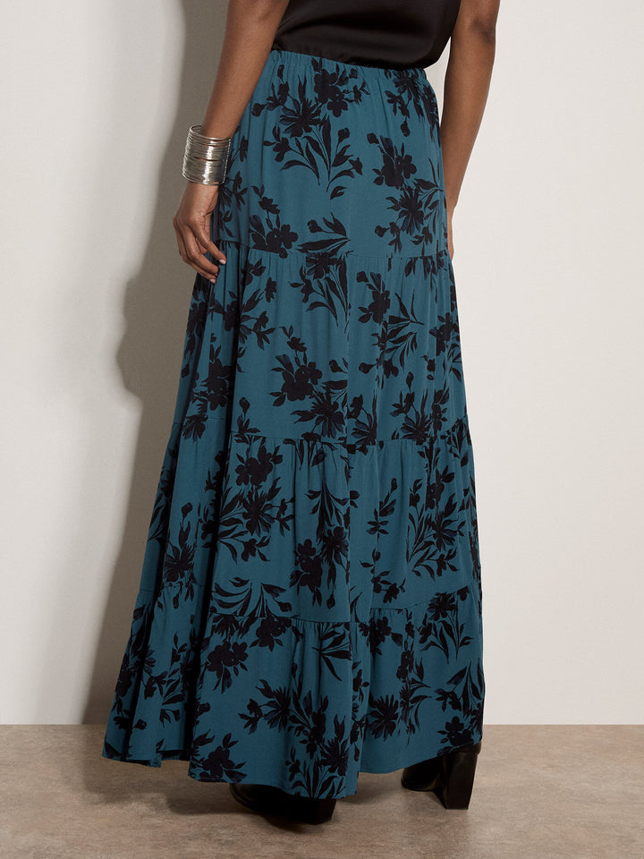A model is shown from the waist down wearing a long teal blue maxi skirt with a black floral pattern, paired with a black top and black heels.