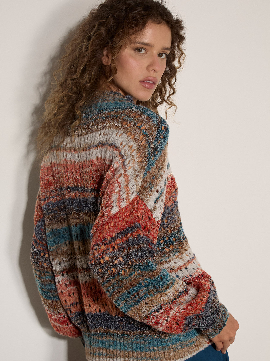 A woman with curly brown hair wearing a chunky, multicolor knit sweater with horizontal stripes of rust red, teal blue, cream, and brown, posing against a plain off-white wall.
