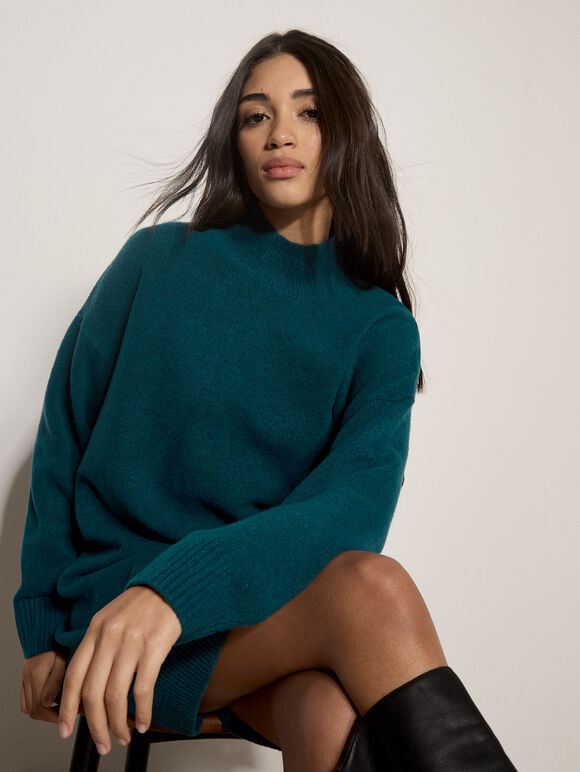 A woman wearing an oversized teal knit sweater sits against a plain light gray wall, with her legs crossed and black knee-high boots visible.