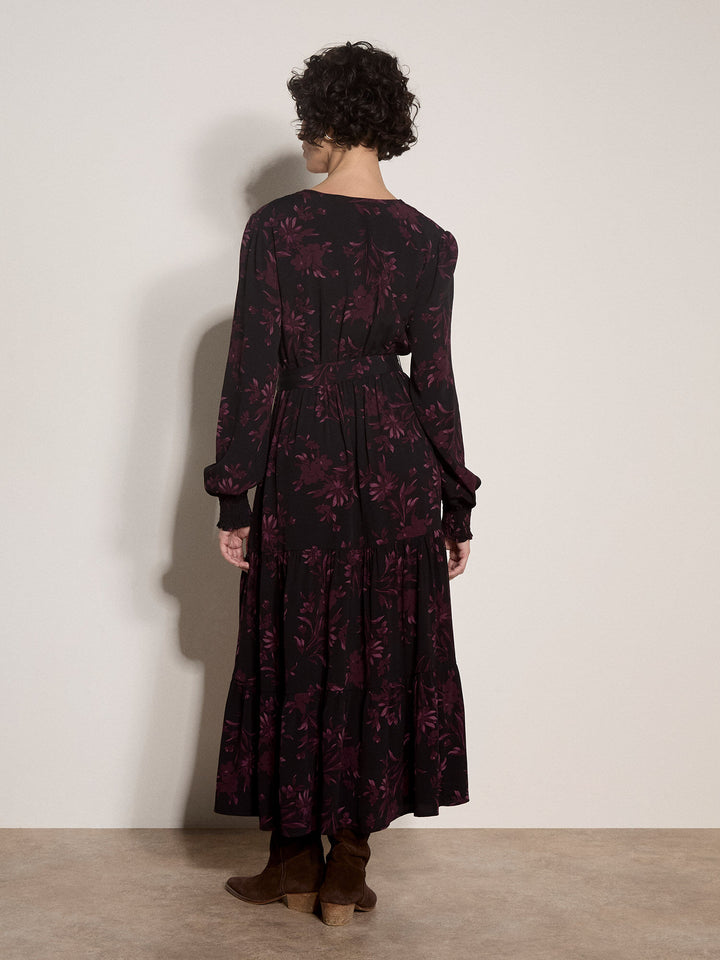 A model viewed from the back wearing a long-sleeved midi dress with a dark floral pattern, paired with brown ankle boots, set against a plain off-white wall.