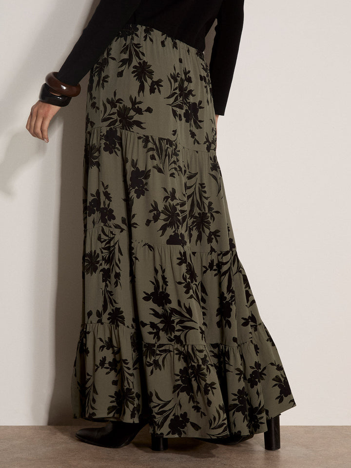 A model is shown from the back wearing a long, tiered maxi skirt in olive green with a black floral pattern, paired with a black top and black heels.