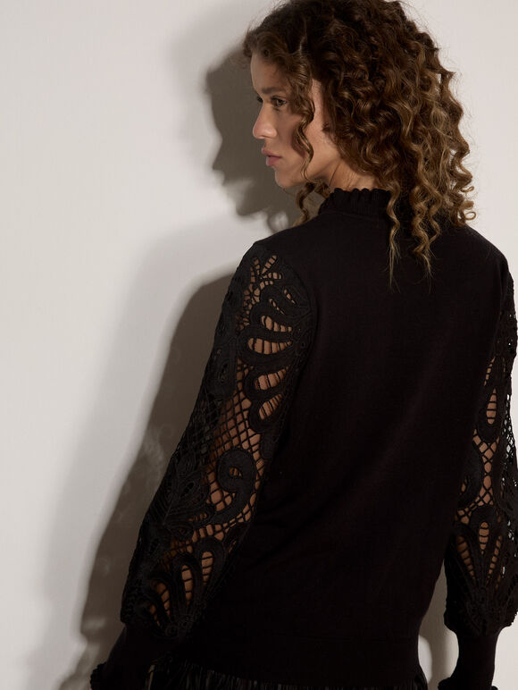 A woman shown from the back wearing a black knit sweater with intricate lace sleeves, posed against a plain white wall with a soft shadow.