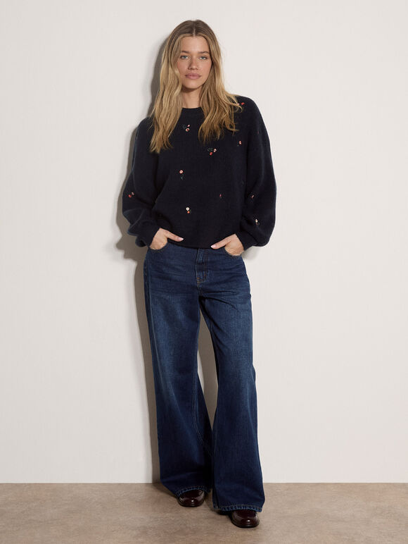 A woman stands against a plain off-white wall wearing a dark navy knit sweater with small metallic embroidered details and dark blue wide-leg jeans.