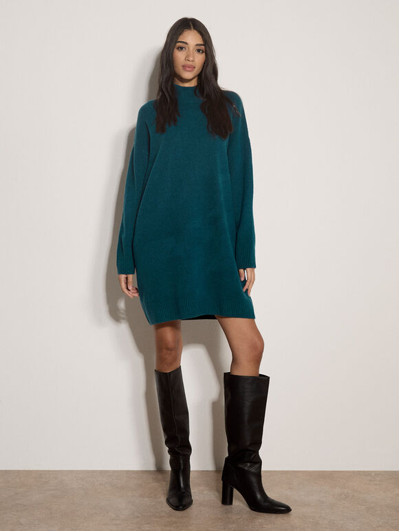 A woman modeling a dark teal green long-sleeve mock-neck knit sweater dress, paired with black knee-high leather boots, standing against a plain off-white wall.