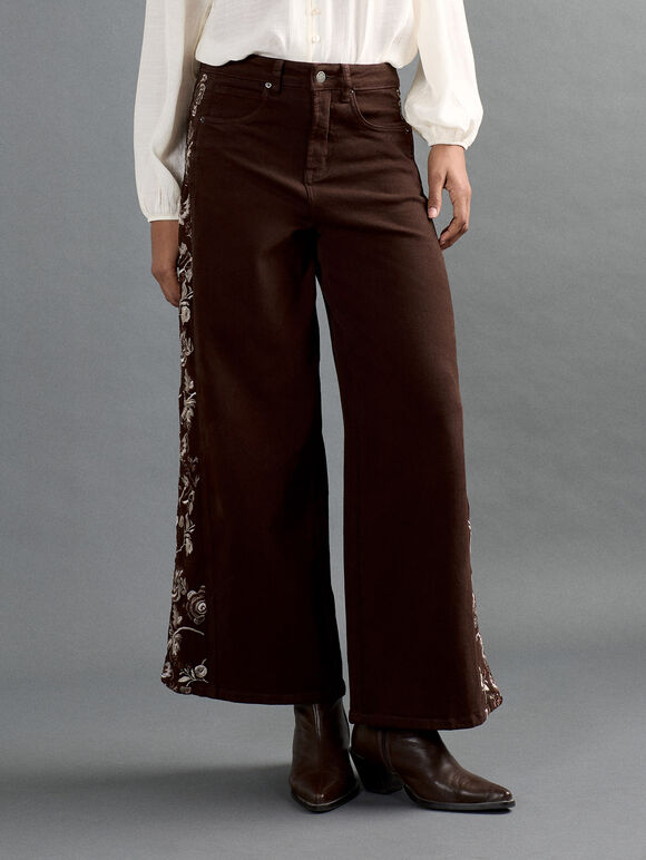 A model is wearing dark brown wide-leg pants with floral embroidery down the side, styled with a white long-sleeve blouse and brown leather boots against a gray studio background.