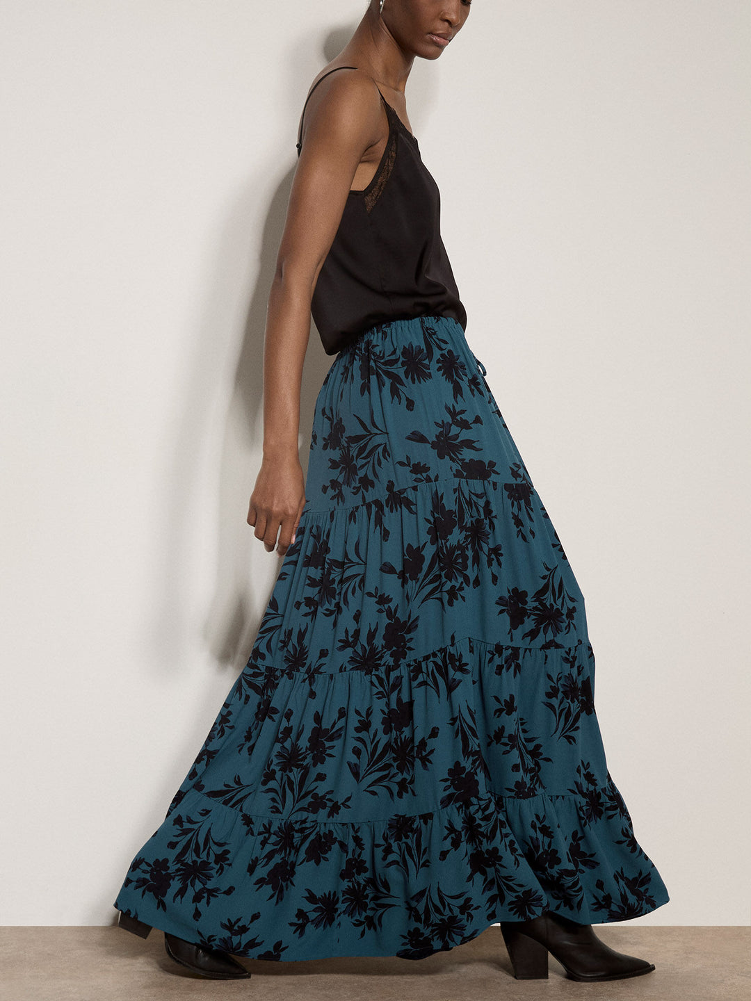 A woman wearing a long teal maxi skirt with a black floral print, paired with a black sleeveless top and black ankle boots, standing against a plain off-white wall.