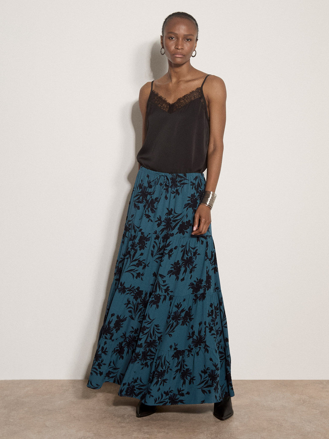 A model stands against a plain off-white wall wearing a dark brown camisole with lace trim and a long teal skirt featuring a black floral pattern.