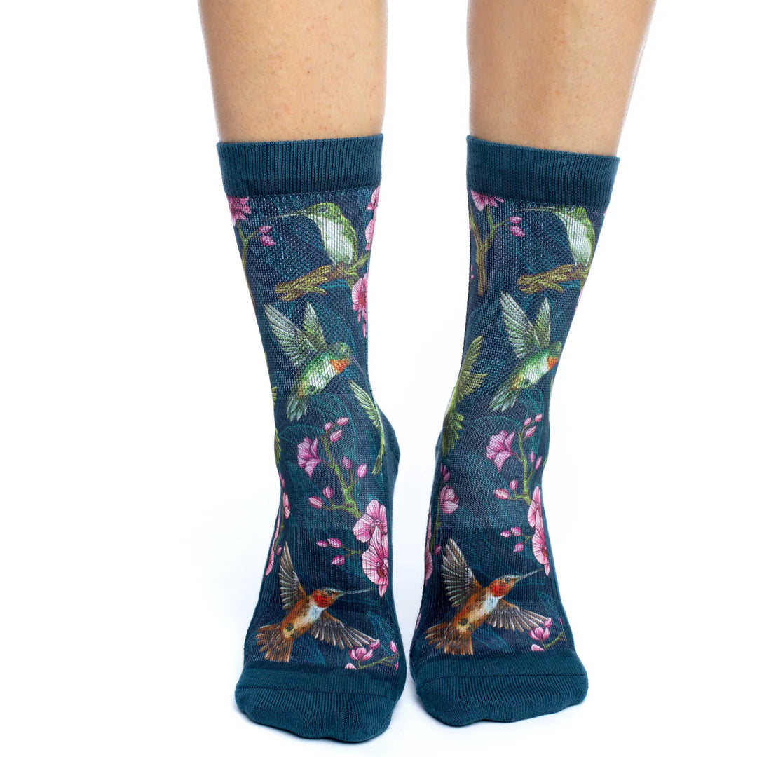 A pair of crew socks worn on feet, featuring a dark teal background with a vibrant print of green hummingbirds, pink flowers, and brown branches.