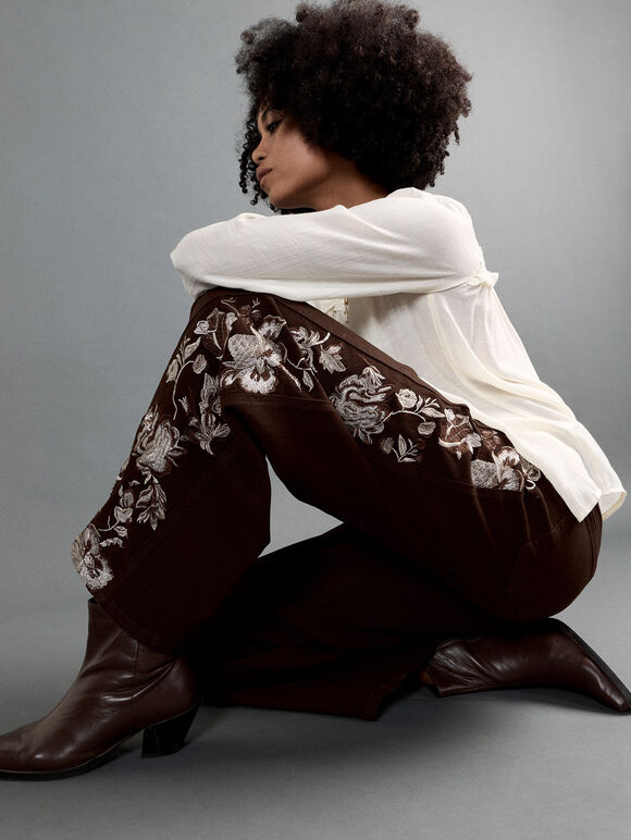 A model with an afro hairstyle is crouching in a studio setting, wearing dark brown wide-leg pants with white floral embroidery, a white long-sleeve top, and dark brown leather boots against a solid gray background.
