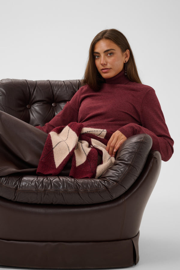 A woman wearing a burgundy turtleneck sweater and dark brown leather pants sits in a dark brown leather armchair, holding a burgundy and beige patterned knit blanket.