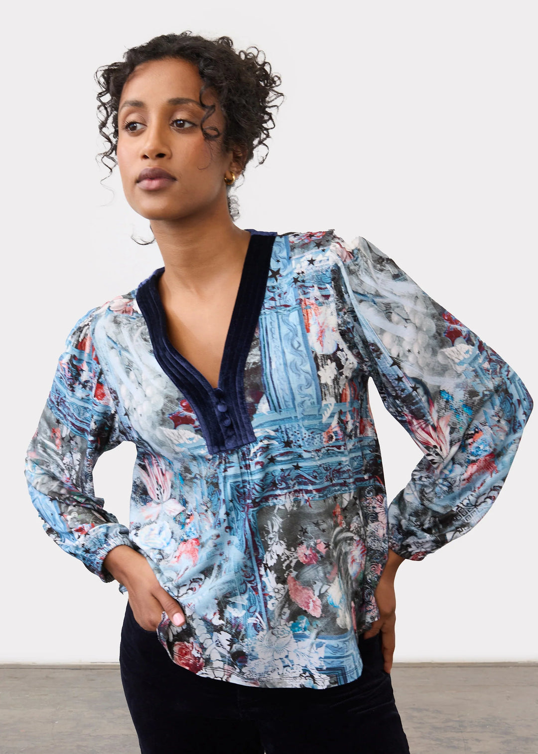 A woman modeling a long-sleeve button-up blouse with a blue, gray, and pink abstract painterly print, featuring a dark navy V-neck trim, paired with black pants against a plain studio background.