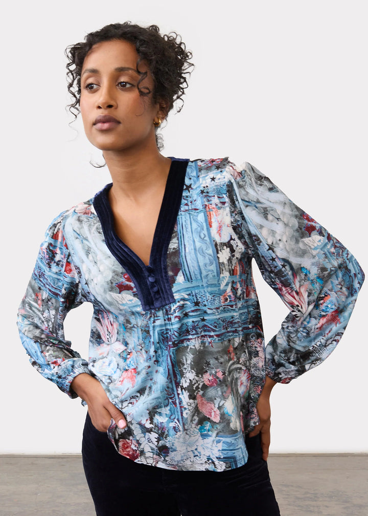A woman modeling a long-sleeve button-up blouse with a blue, gray, and pink abstract painterly print, featuring a dark navy V-neck trim, paired with black pants against a plain studio background.