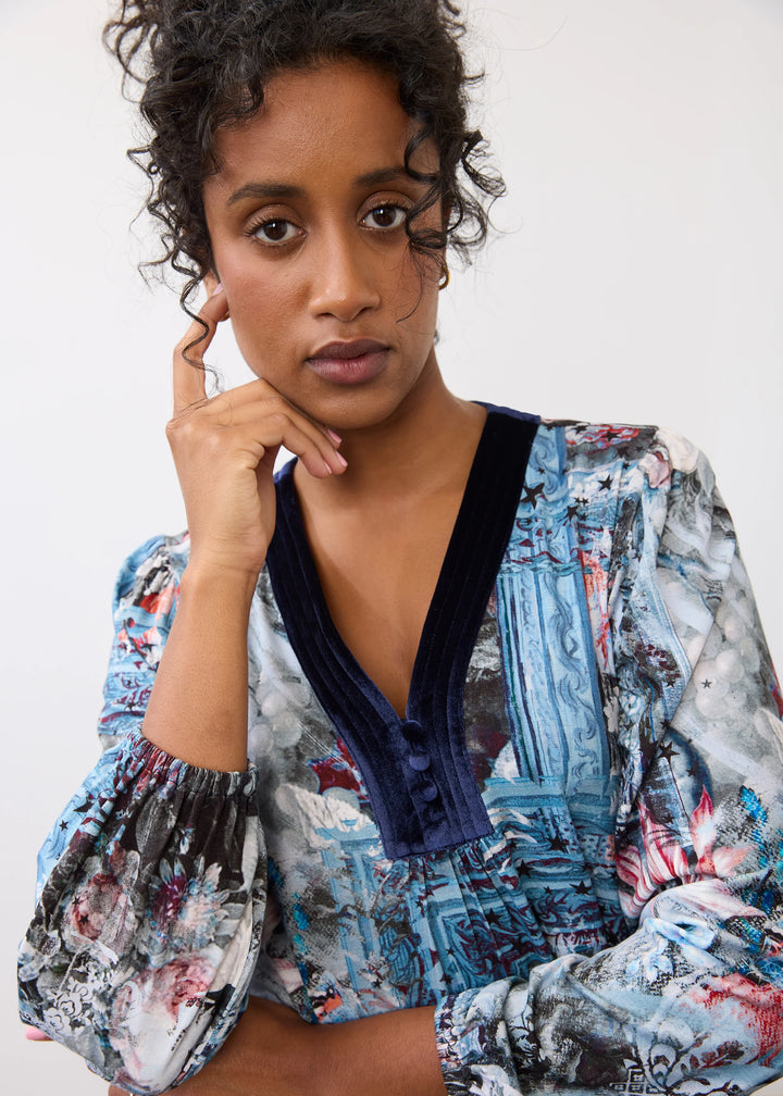 A woman wearing a long-sleeve blouse with a blue and gray architectural floral print, featuring a dark navy velvet V-neckline, posing against a plain white background.