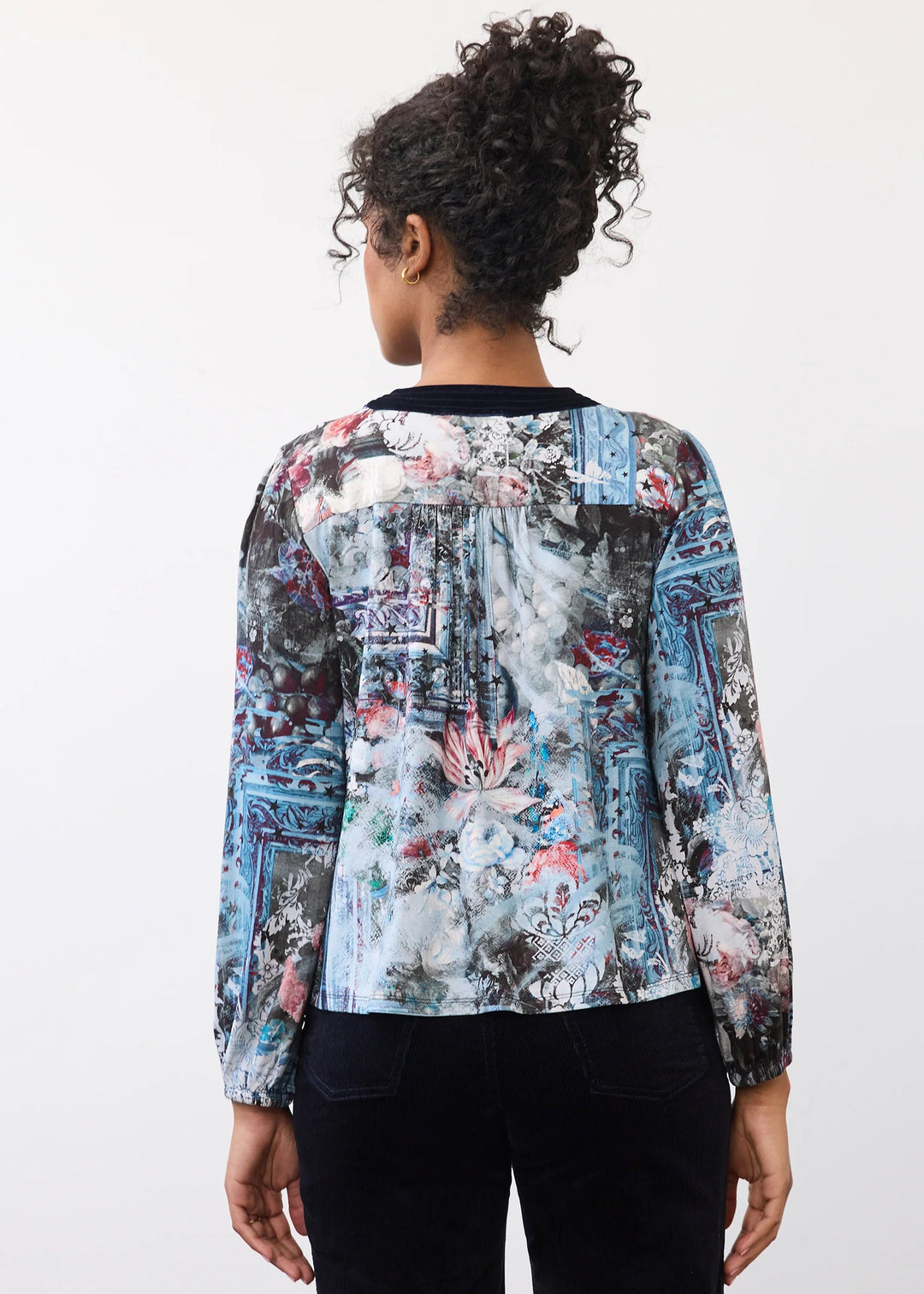 A woman viewed from behind wearing a long-sleeve blouse with a blue, gray, and pink abstract floral print, paired with black pants, against a plain white background.