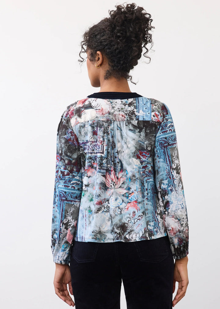 A woman viewed from behind wearing a long-sleeve blouse with a blue, gray, and pink abstract floral print, paired with black pants, against a plain white background.
