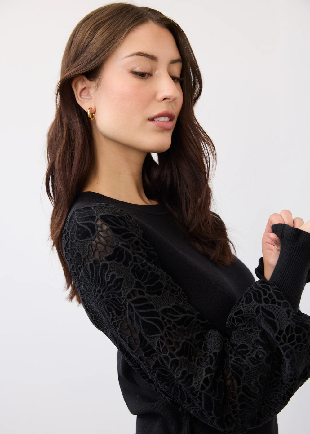 A woman wearing a black long-sleeve top with intricate floral lace applique on the sleeves, shown against a plain light gray studio background.