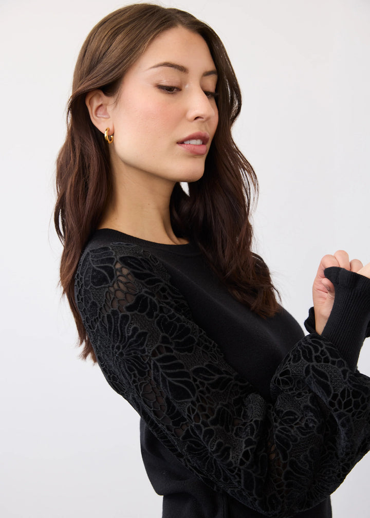 A woman wearing a black long-sleeve top with intricate floral lace applique on the sleeves, shown against a plain light gray studio background.