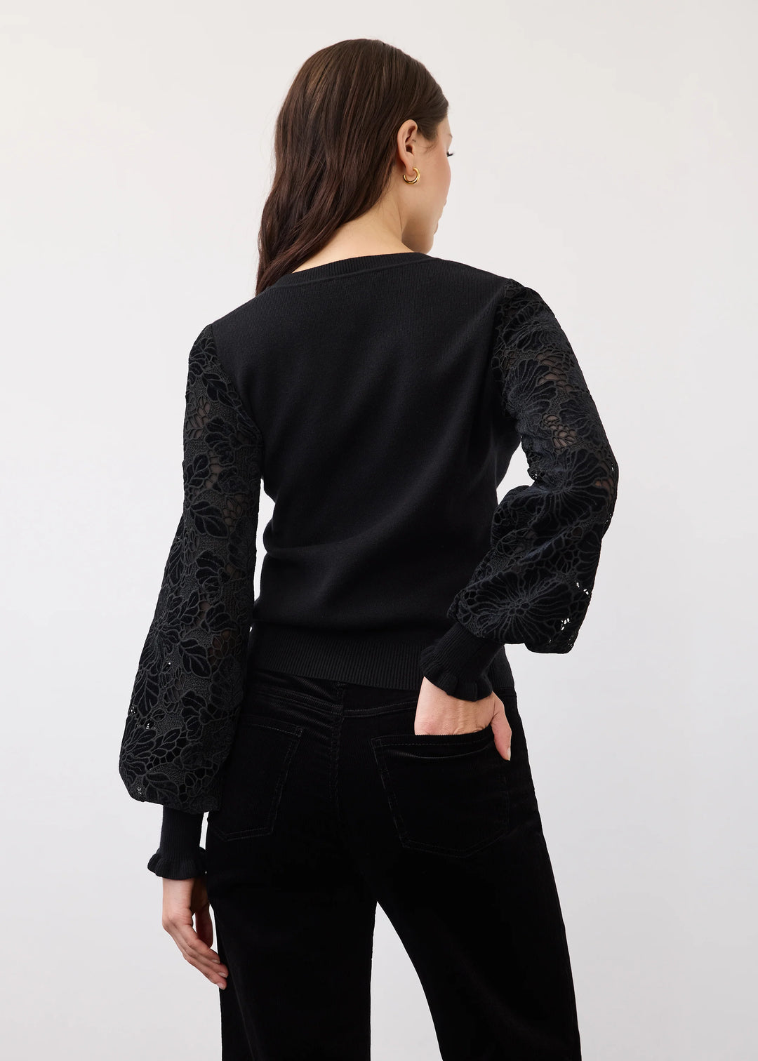 A woman shown from the back wearing a black long-sleeve top with sheer floral lace sleeves and black pants, photographed against a plain light gray studio background.