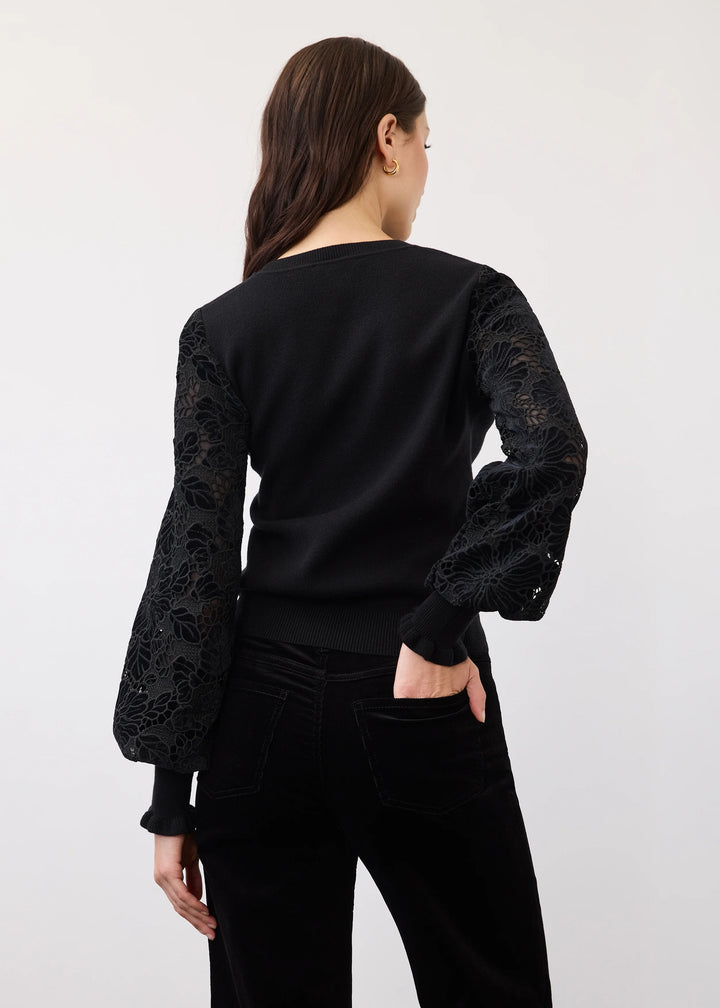 A woman shown from the back wearing a black long-sleeve top with sheer floral lace sleeves and black pants, photographed against a plain light gray studio background.