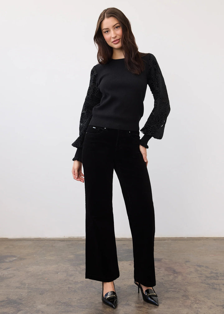A woman modeling a black long-sleeve knit top with textured sleeves and matching black wide-leg trousers, paired with black pointed-toe heels, standing against a plain white wall.