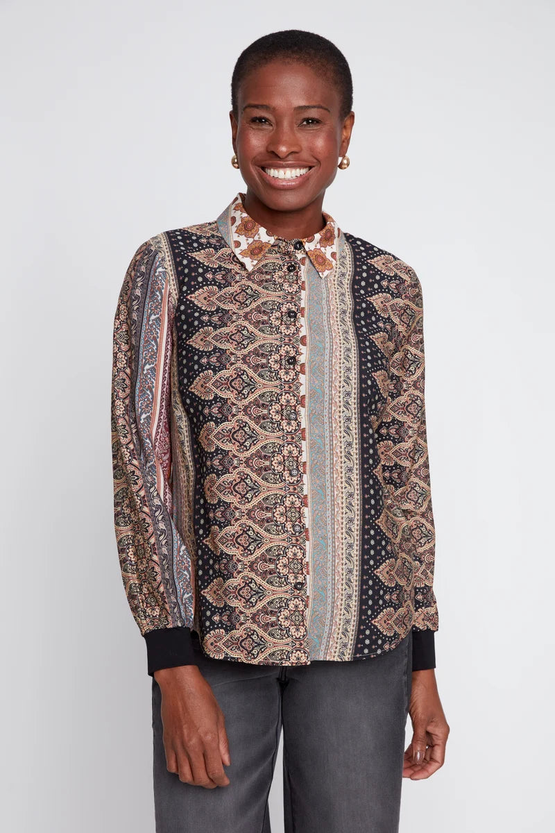 A smiling woman wearing a long-sleeve button-up blouse with a complex geometric and tribal pattern in beige, brown, black, and muted blue, paired with gray trousers, against a light gray studio background.
