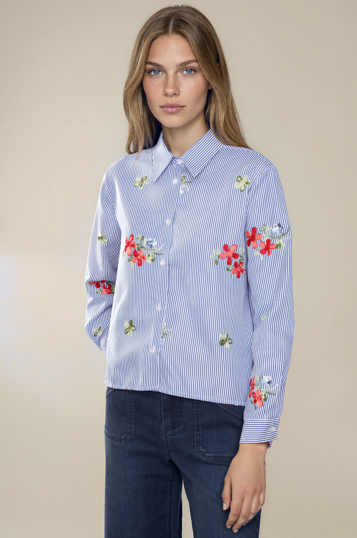 A woman modeling a light blue and white striped long-sleeve button-up shirt with red and green floral embroidery, paired with dark blue jeans, against a solid beige background.