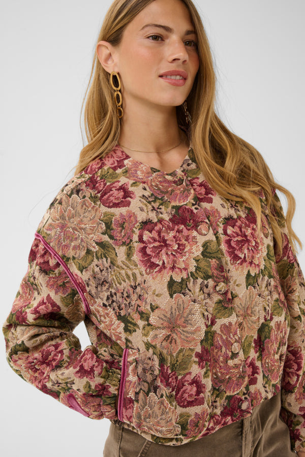 A woman wearing a cropped, long-sleeve floral jacket with a vintage floral pattern in pink, green, and beige, paired with olive pants against a light gray background.