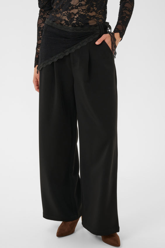 A model wearing black wide-leg trousers with a draped wrap-style top, paired with a black lace long-sleeve top and brown pointed-toe heels, photographed against a light gray studio background.
