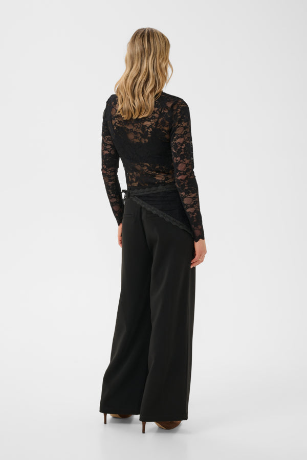 A woman shown from the back wearing a black lace long-sleeve top and black wide-leg trousers against a plain light gray studio background.