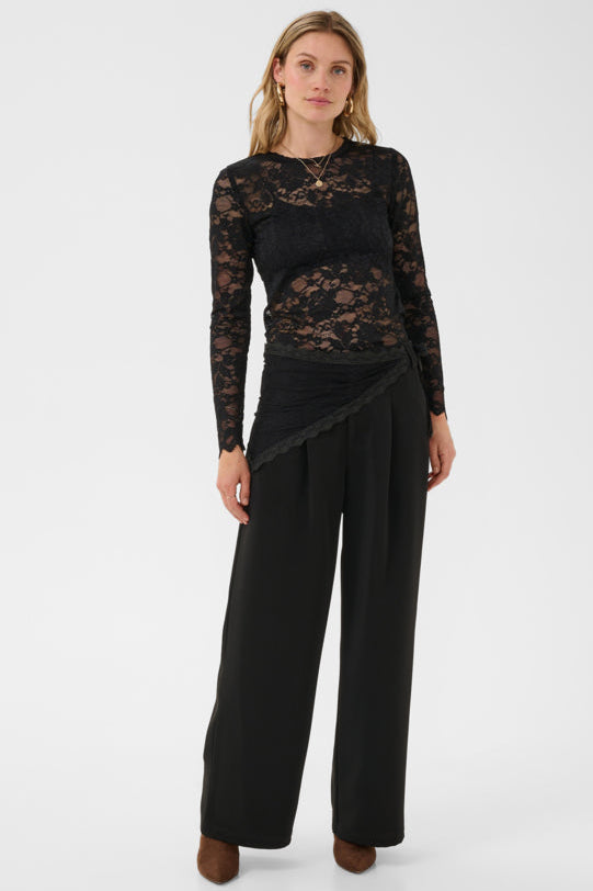 A woman modeling a black long-sleeve top with a sheer floral lace bodice and wide-leg black trousers, standing against a plain light gray studio background.
