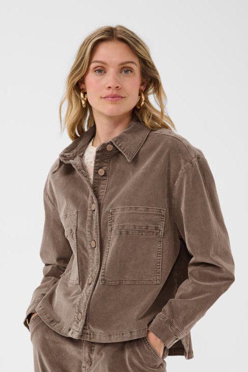 A woman wearing a brown corduroy button-up shirt jacket and matching corduroy pants, standing against a light gray studio background.