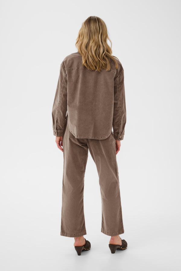 A woman viewed from the back wearing a matching brown corduroy long-sleeve shirt and wide-leg trousers, paired with black mule heels, against a plain light gray studio background.