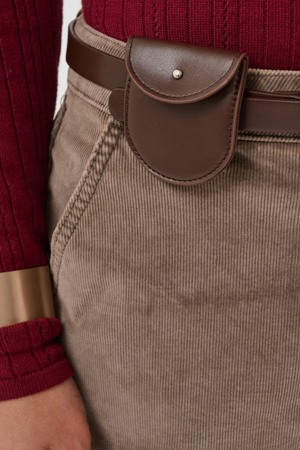 A close-up of a person wearing a brown leather belt with a small flap pouch and a silver stud, paired with taupe corduroy trousers and a burgundy ribbed sweater.