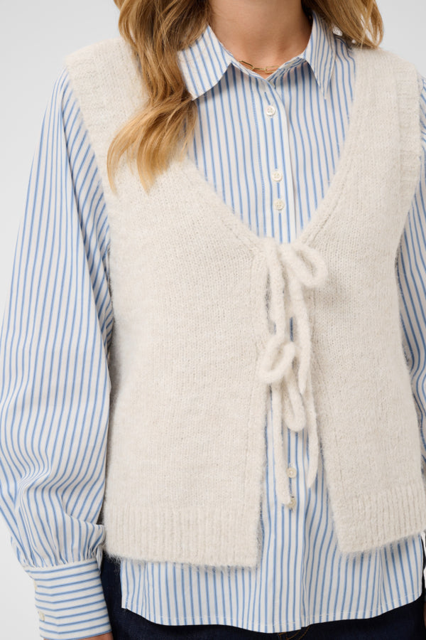 A woman wearing a cream-colored, fuzzy knit cardigan vest over a light blue and white striped button-down shirt.