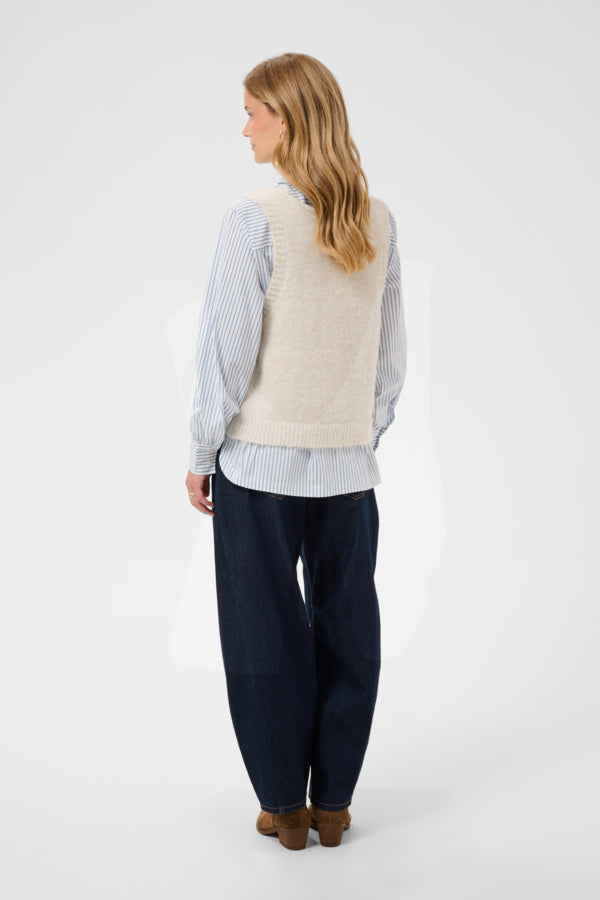 A woman is shown from the back wearing a cream-colored knit vest over a light blue and white striped shirt, paired with dark navy wide-leg pants and brown shoes, against a plain light gray studio background.