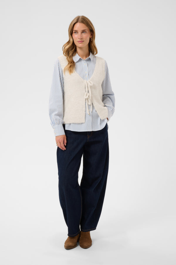 A woman stands against a light gray studio background wearing a cream knit vest over a light blue and white striped button-up shirt, dark navy wide-leg pants, and brown ankle boots.
